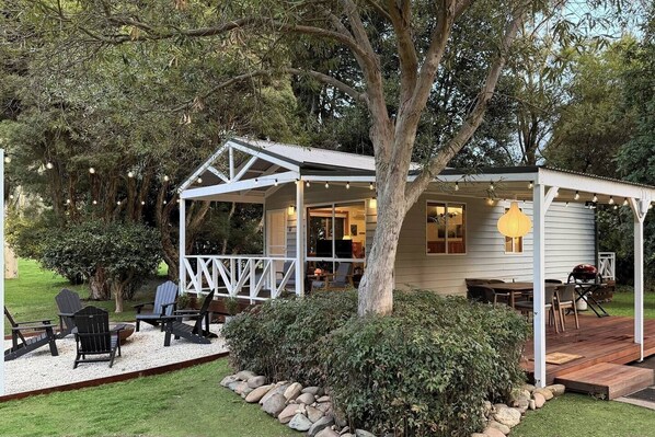 A covered outdoor seating area with a table and chairs, surrounded by a garden and a house.