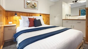 A hotel room with a wooden headboard, a bed with white bedding and multiple pillows, a nightstand, a dresser, a mirror, and a framed artwork on the wall.