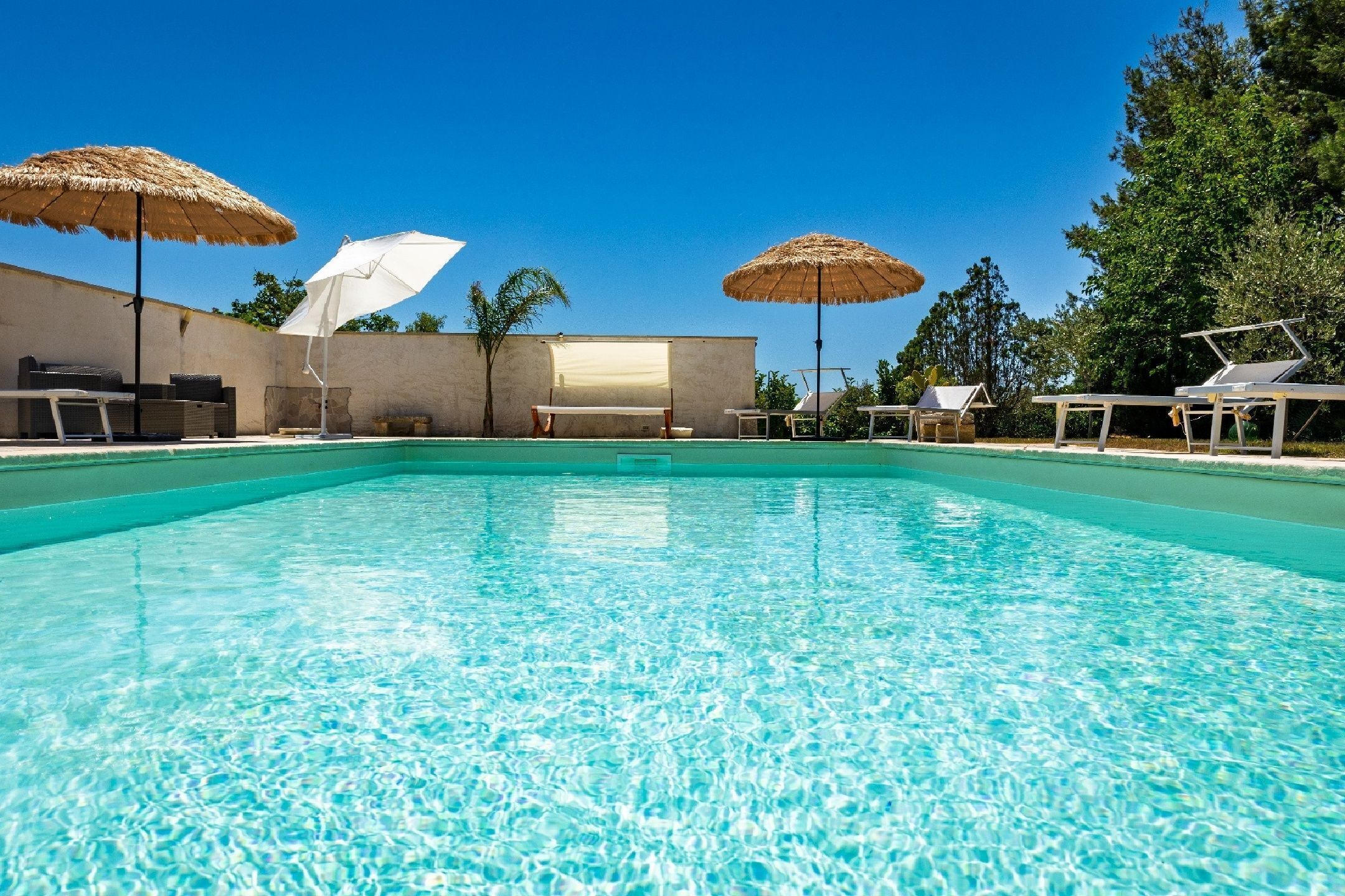 Una piscina con lettini e ombrelloni, circondata da un muretto e alberi.