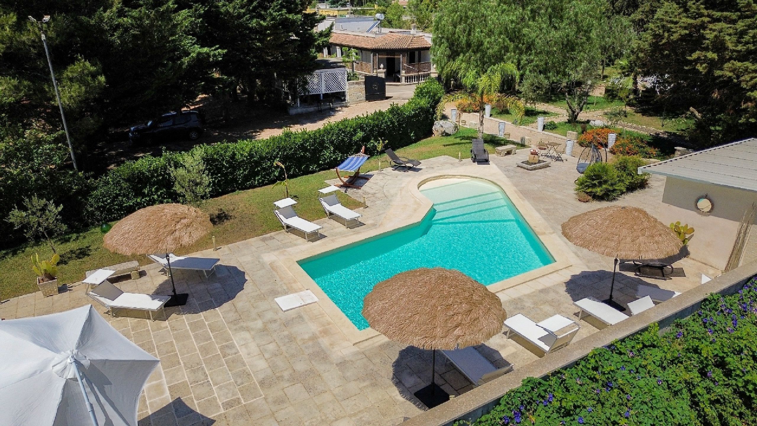 Une piscine entourée de chaises longues et de parasols, elle-même bordée d’un jardin aménagé et d’un bâtiment en arrière-plan.
