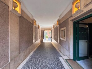Ein Korridor mit gefliestem Boden, einer grünen Tür rechts und Blick in einen Raum mit Bett und Fenster.