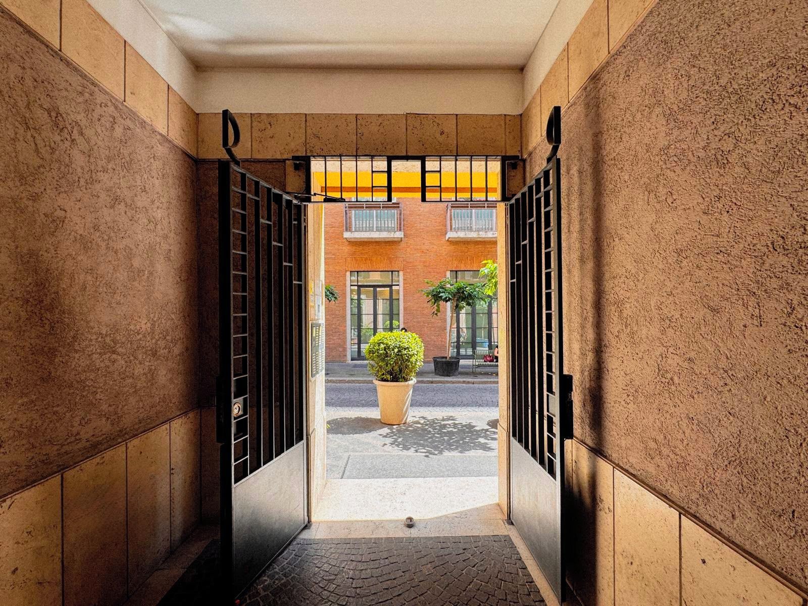 Une étroite entrée, fermée par un portail en métal, mène à un bâtiment dont la façade est en briques et devant lequel se trouve un pot de fleurs.