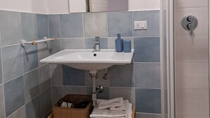Ein Badezimmer mit Waschbecken in Weiß, blauen Fliesen, einem Spiegel, einem Holzregal mit Handtüchern und einem Mülleimer.