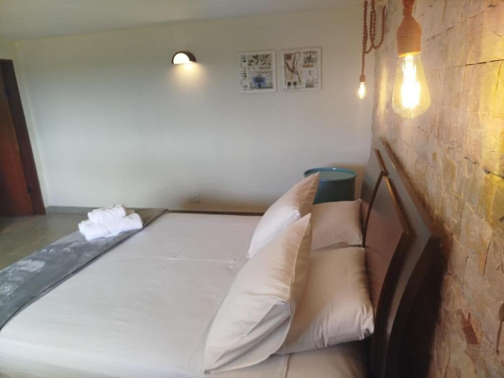Una habitación de hotel con una cama, almohadas, un basurero azul y lámparas fijadas a la pared.