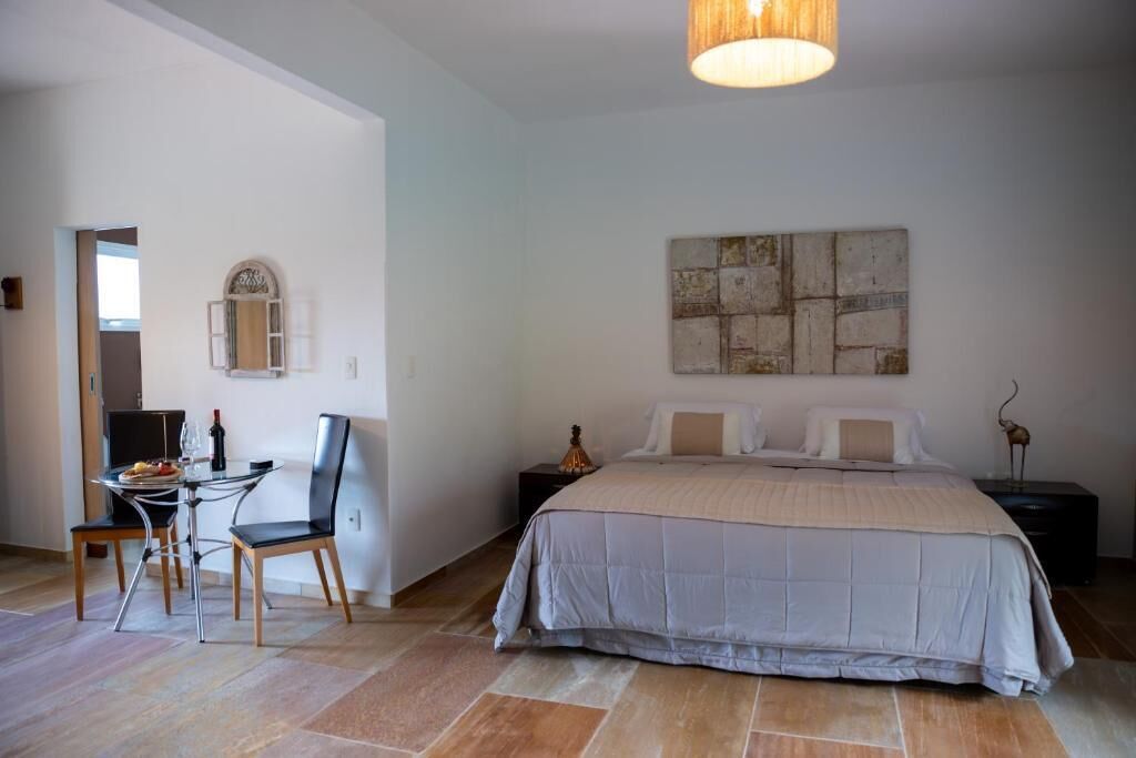 A bedroom with a large bed, a bedside table, a nightstand, a wall-mounted artwork, and a dining area with a table and chairs.