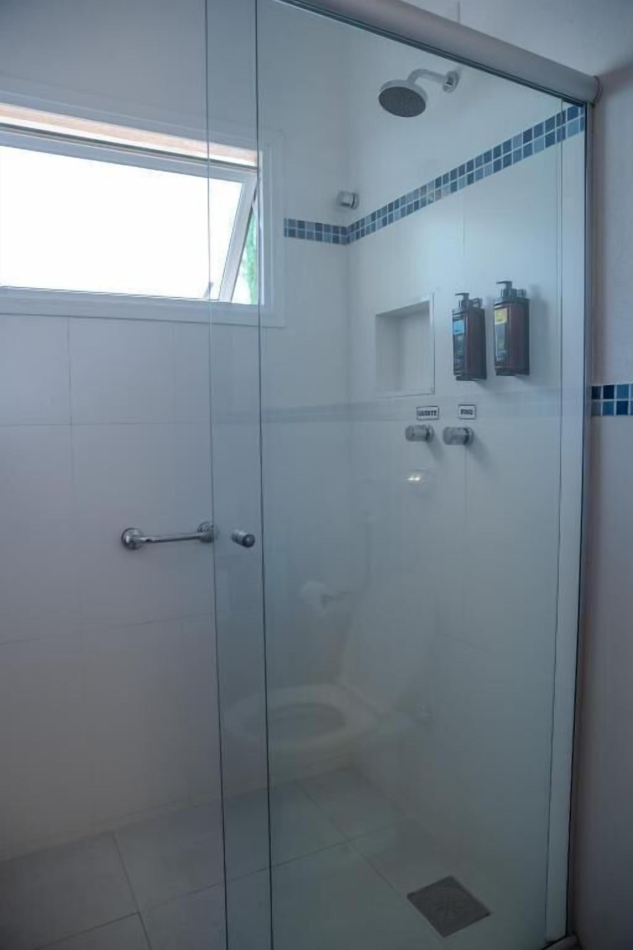 A glass-enclosed shower stall with a toilet visible through the glass, a showerhead above, and a small shelf with toiletries.