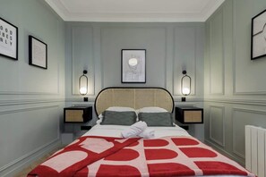 Una cama bien hecha con colcha de cuadros rojos y blancos, dos mesitas de noche con lámparas y cuadros en las paredes.
