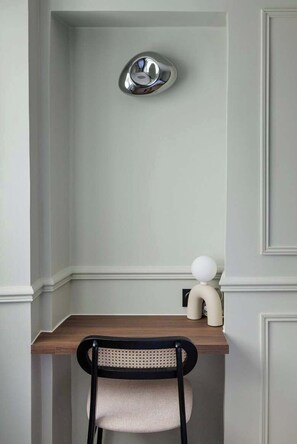 Un coin de pièce avec un mur de couleur claire, un bureau en bois, une chaise noire avec un siège en osier, et un petit objet blanc accroché au mur.
