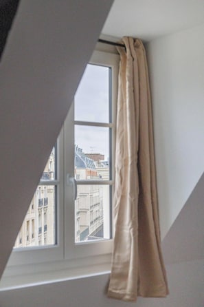 A window with a curtain, a view of buildings, and a sloped ceiling. - Charming Flat near Madeleine (Paris)