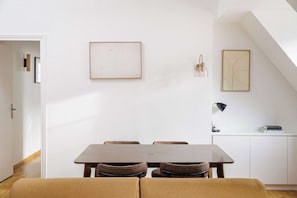 A dining area with a table and chairs, a white wall with two framed artworks, a lamp, and a white cabinet with a book on top. - Charming Flat near Madeleine (Paris)