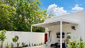 Un espacio al aire libre techado con equipo de ejercicio, una pared blanca y un árbol.