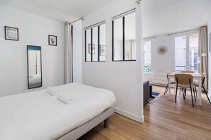 A hotel room with a bed, a desk, and a chair. There are windows and a view of the outside. - Greneta - Spacious Flat Montorgueuil 4PAX (Paris)
