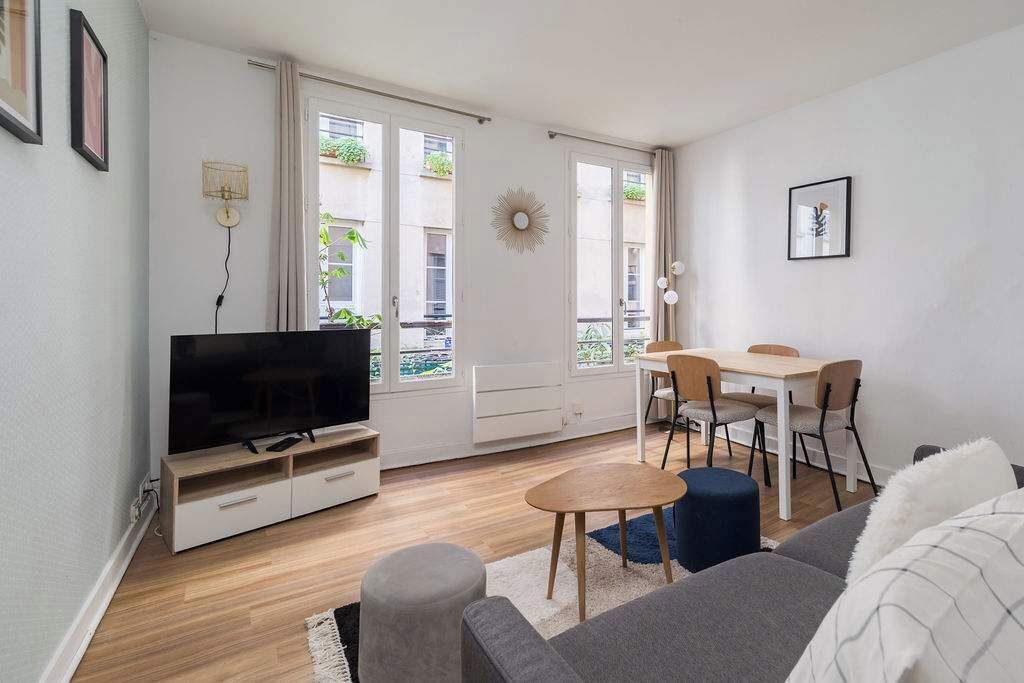 A living room with a TV, a dining table with chairs, a sofa, and a window with plants.
