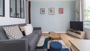 A modern living room with a grey sofa, a small wooden coffee table, a blue ottoman, a flat-screen TV on a wooden stand, and a large window with curtains. - Greneta - Spacious Flat Montorgueuil 4PAX (Paris)