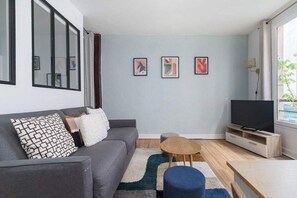 A modern living room with a grey sofa, a small wooden coffee table, a blue ottoman, a flat-screen TV on a wooden stand, and a large window with curtains. - Greneta - Spacious Flat Montorgueuil 4PAX (Paris)