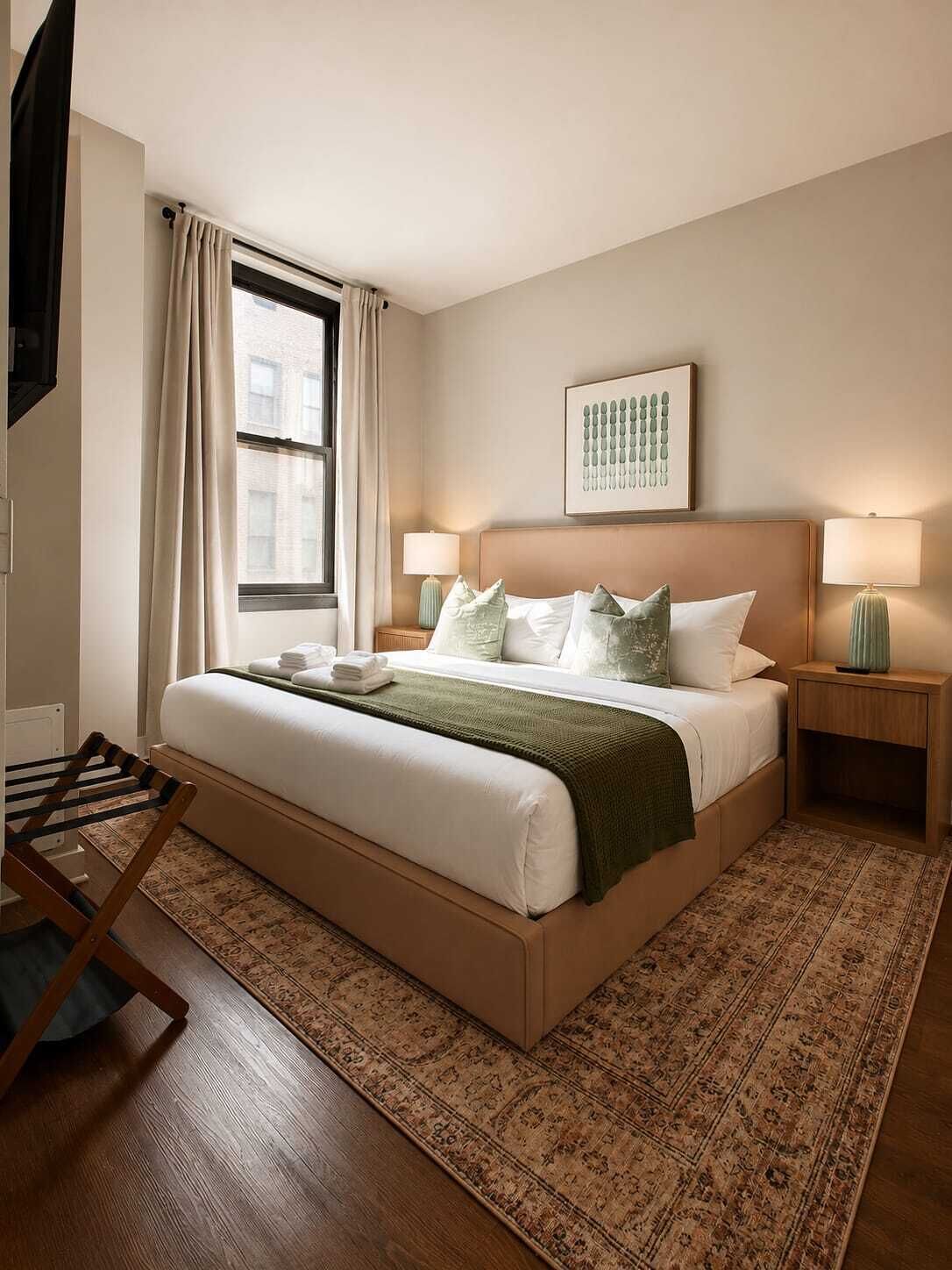 A bedroom with a large bed, two bedside tables, a window with curtains, a wall-mounted artwork, and a patterned rug.