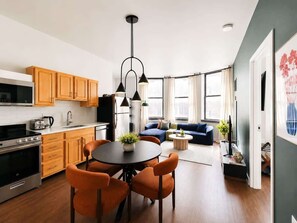 A modern kitchen with wooden cabinets, a dining area with orange chairs, and a living room with a blue sofa. - Modern 2BR in Heart of Philly | Rafa Properties (Philadelphia)