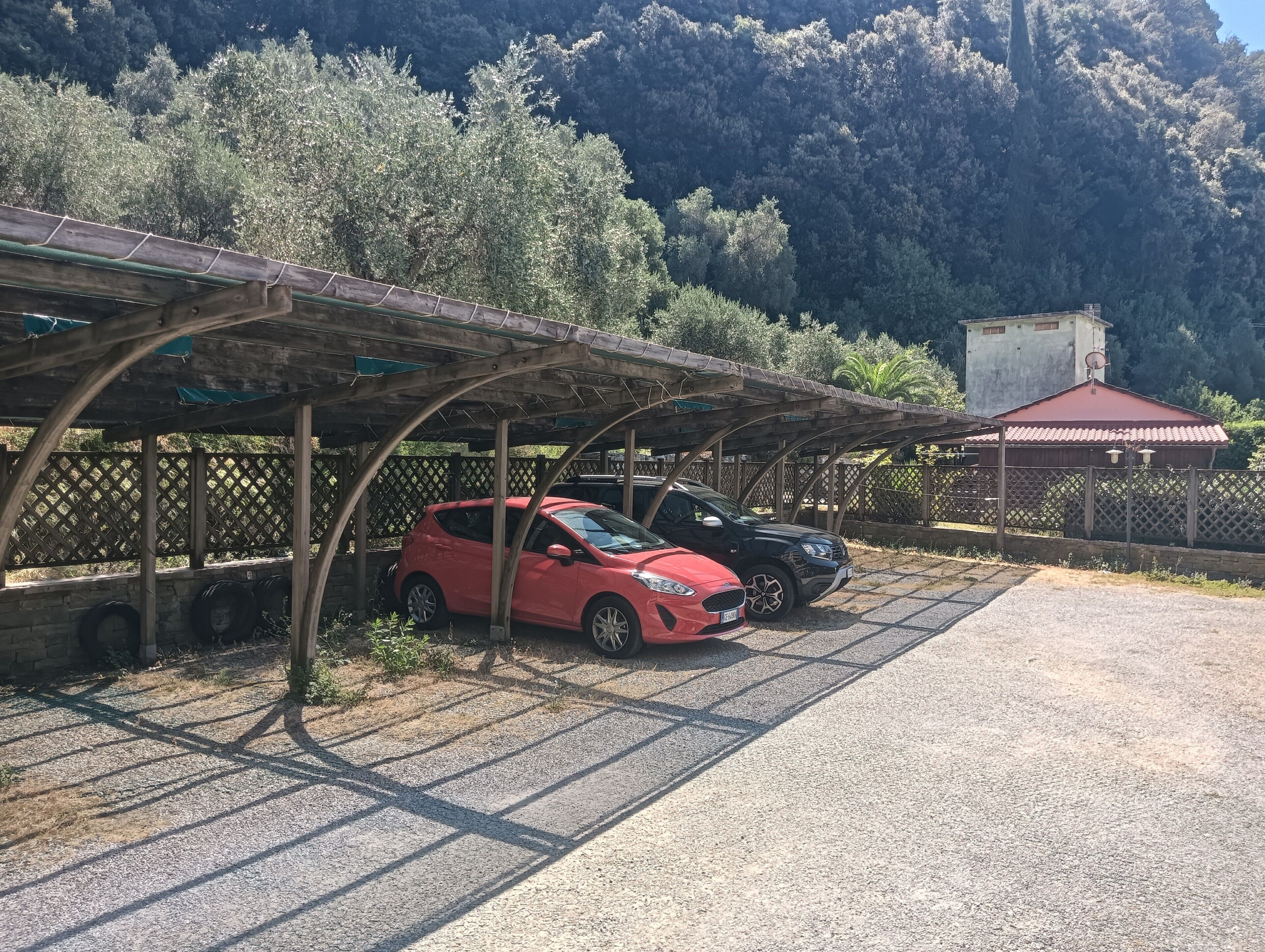 Een overdekte parkeerplaats met twee auto's, een houten constructie en een gebouw op de achtergrond.