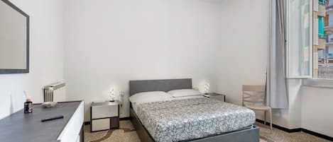 A bedroom with a bed, bedside tables, a dresser, a radiator, and a window with a view of buildings.