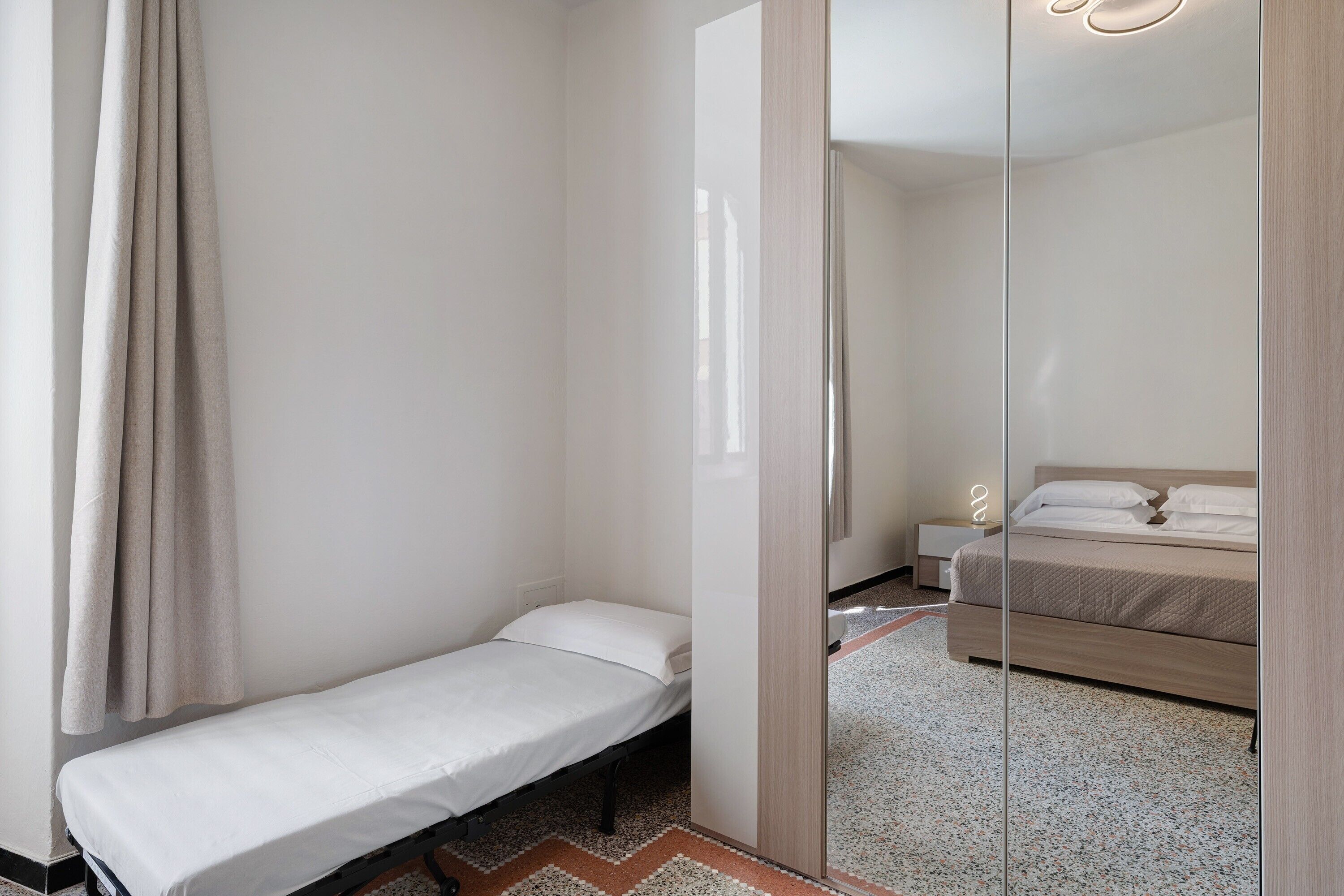 A small, single-bed room with a wooden floor, a bedside table, and a mirror.