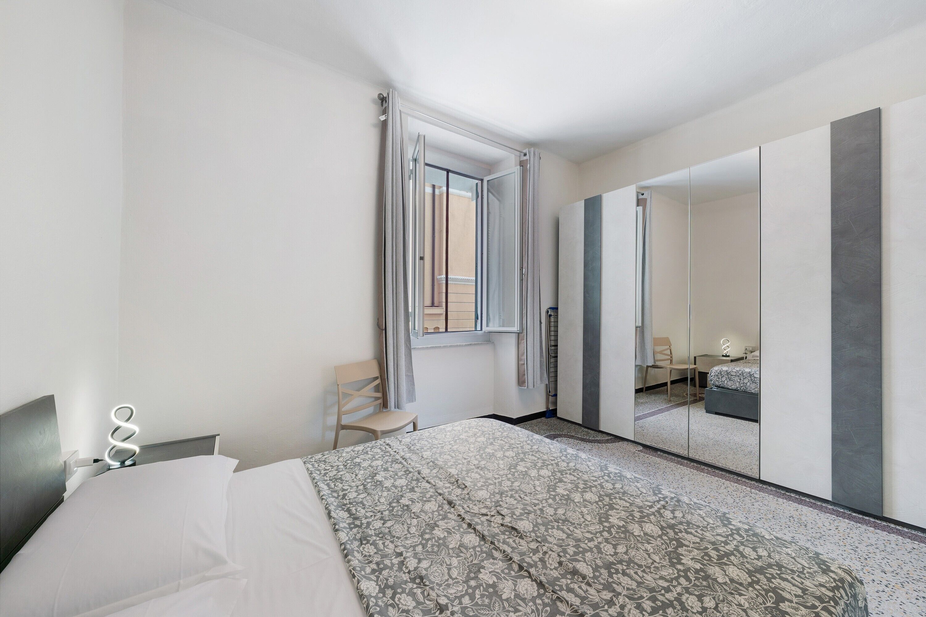Una camera d'albergo con un letto, una sedia, uno specchio e una finestra con tende.