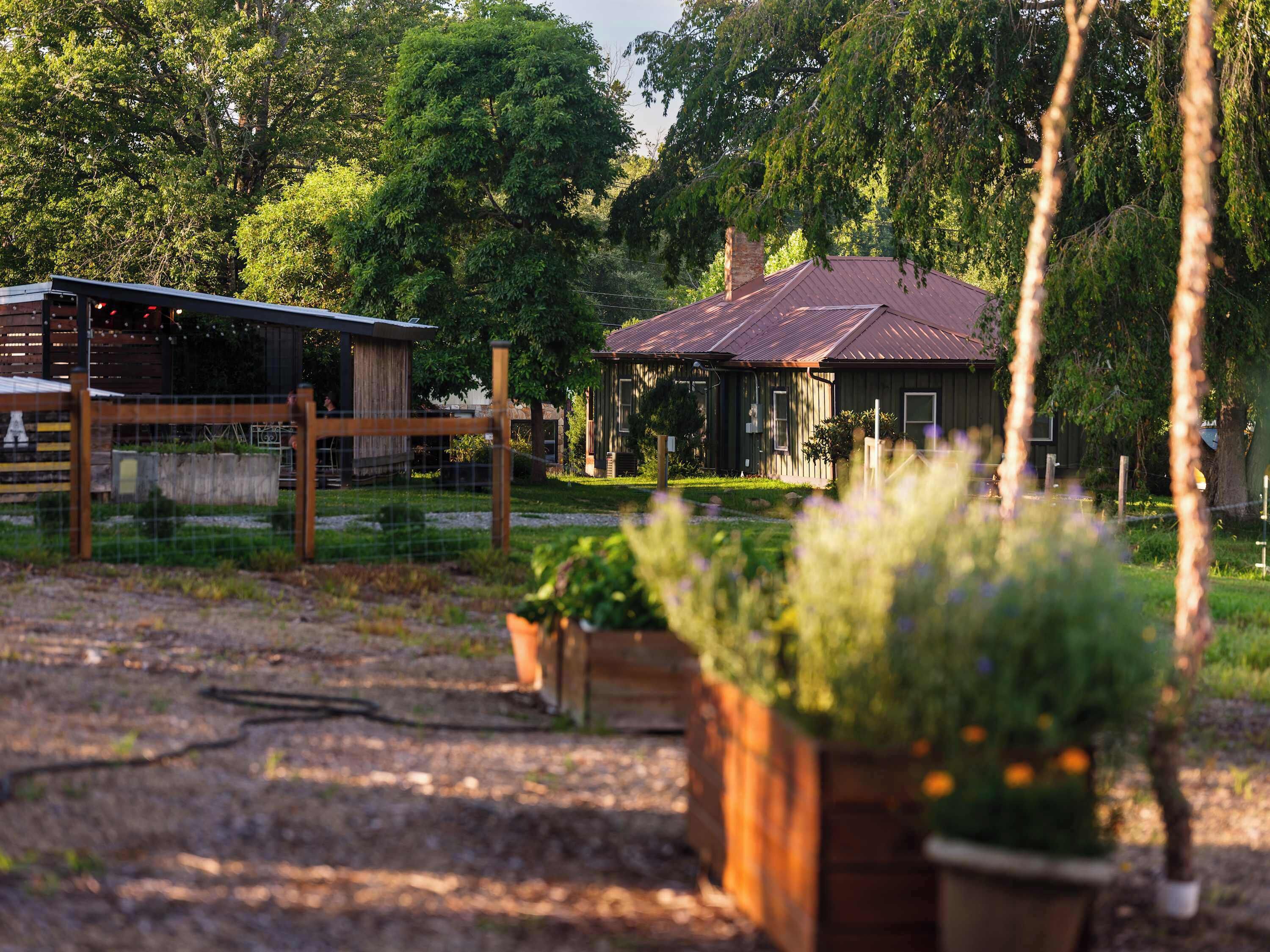 A rustic building with a metal roof, surrounded by greenery and a fenced area.