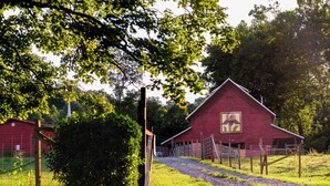 Ein rotes Bauernhaus mit einem auffälligen, geometrischen Logo an der Seite, umgeben von üppigem Grün und einem Kiesweg, der dorthin führt.
