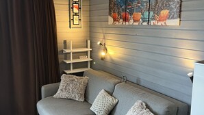 Een grijze bank met drie kussens, een houten bijzettafeltje, een wandplank met ingekaderde foto's en een schilderij van een besneeuwd landschap.