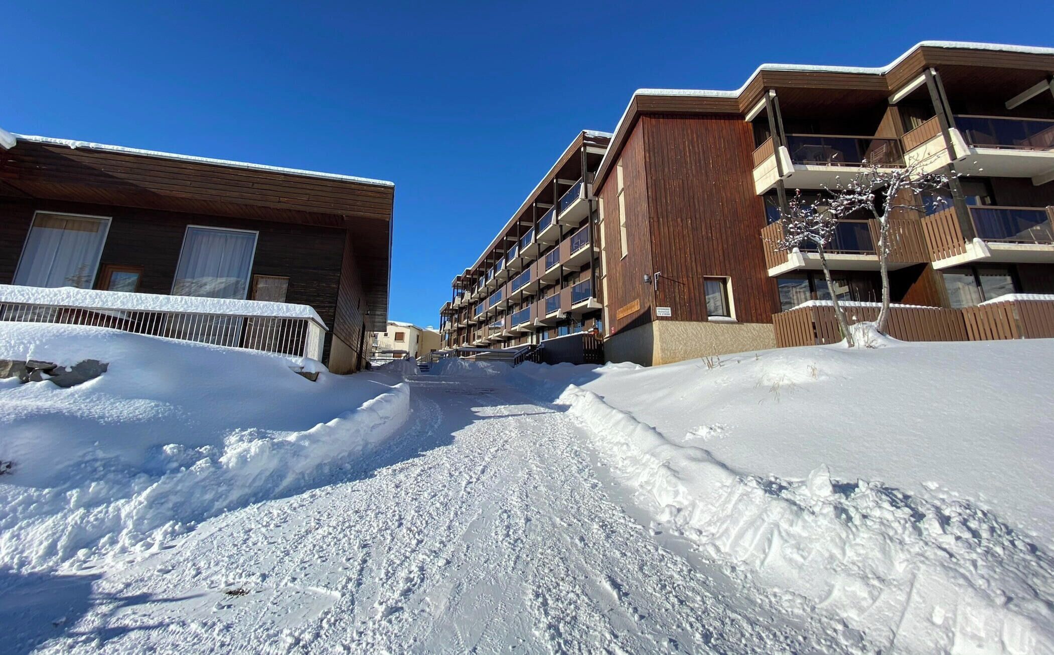 A snow-covered street with modern buildings, including a multi-story structure with balconies.