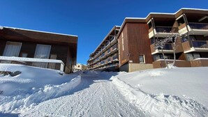 A snow-covered street with modern buildings, including a multi-story structure with balconies.
