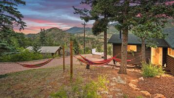 A rustic cabin with a hammock, surrounded by trees and a view of a mountainous landscape.