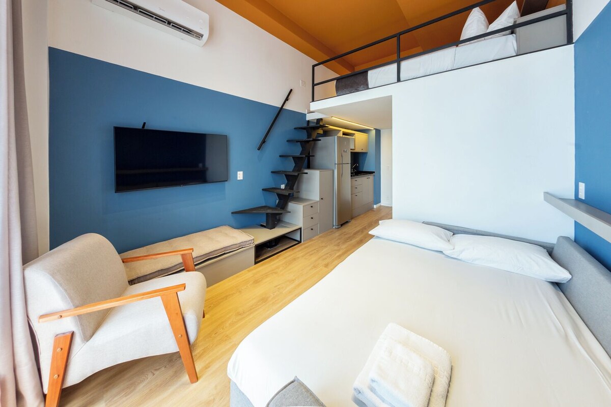 A modern hotel room with a bed, a desk, a chair, a TV, and a kitchenette.