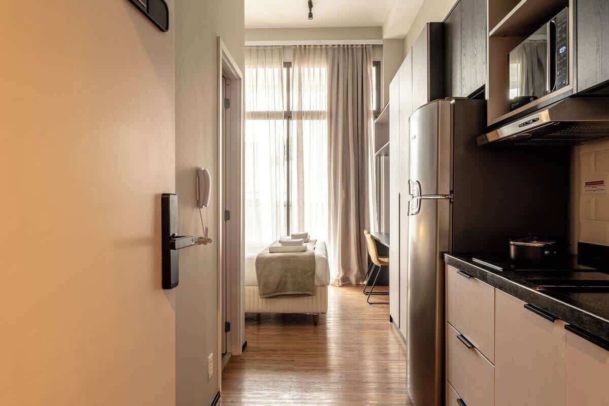 A narrow hallway leading to a bedroom with a bed, a kitchenette with a refrigerator and microwave, and a window with curtains.