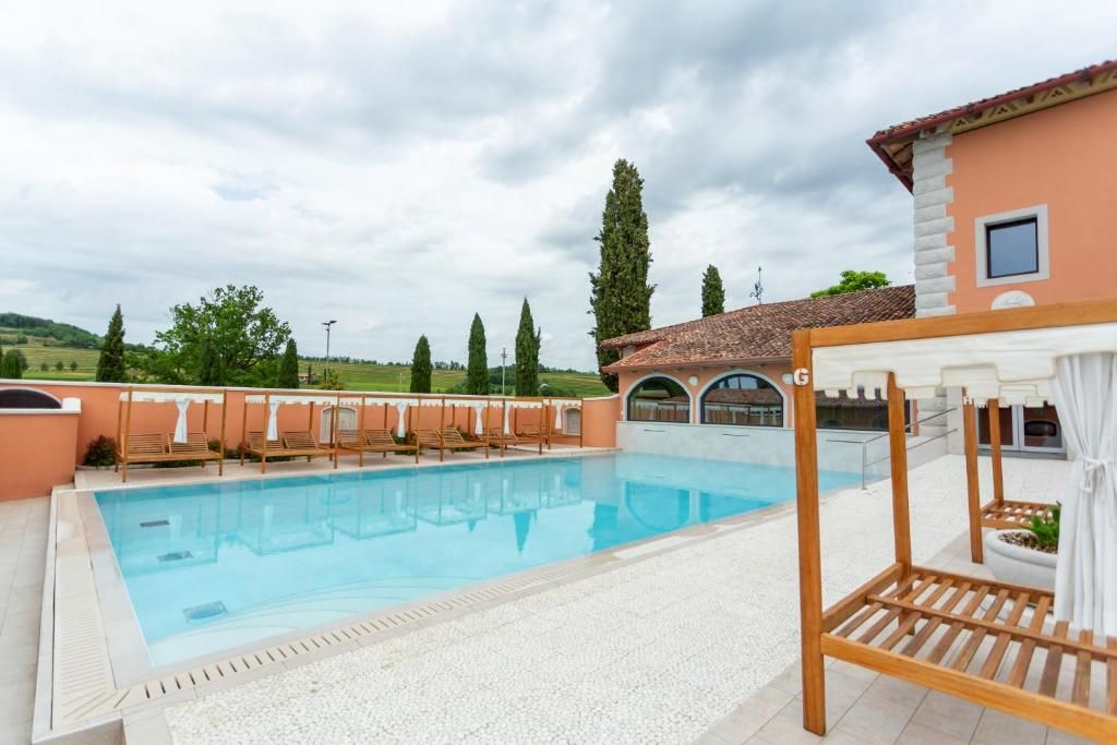 Una piscina con lettini prendisole e un pergolato, circondata da un edificio con tetto in tegole e vista sulla campagna.