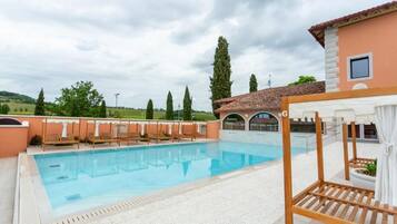 Una piscina con lettini prendisole e un pergolato, circondata da un edificio con tetto in tegole e vista sulla campagna.