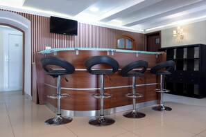 A reception area with a wooden counter, four bar stools, a flat-screen TV, and a chandelier. - Villa Oliver Abádszalók (Abádszalók)