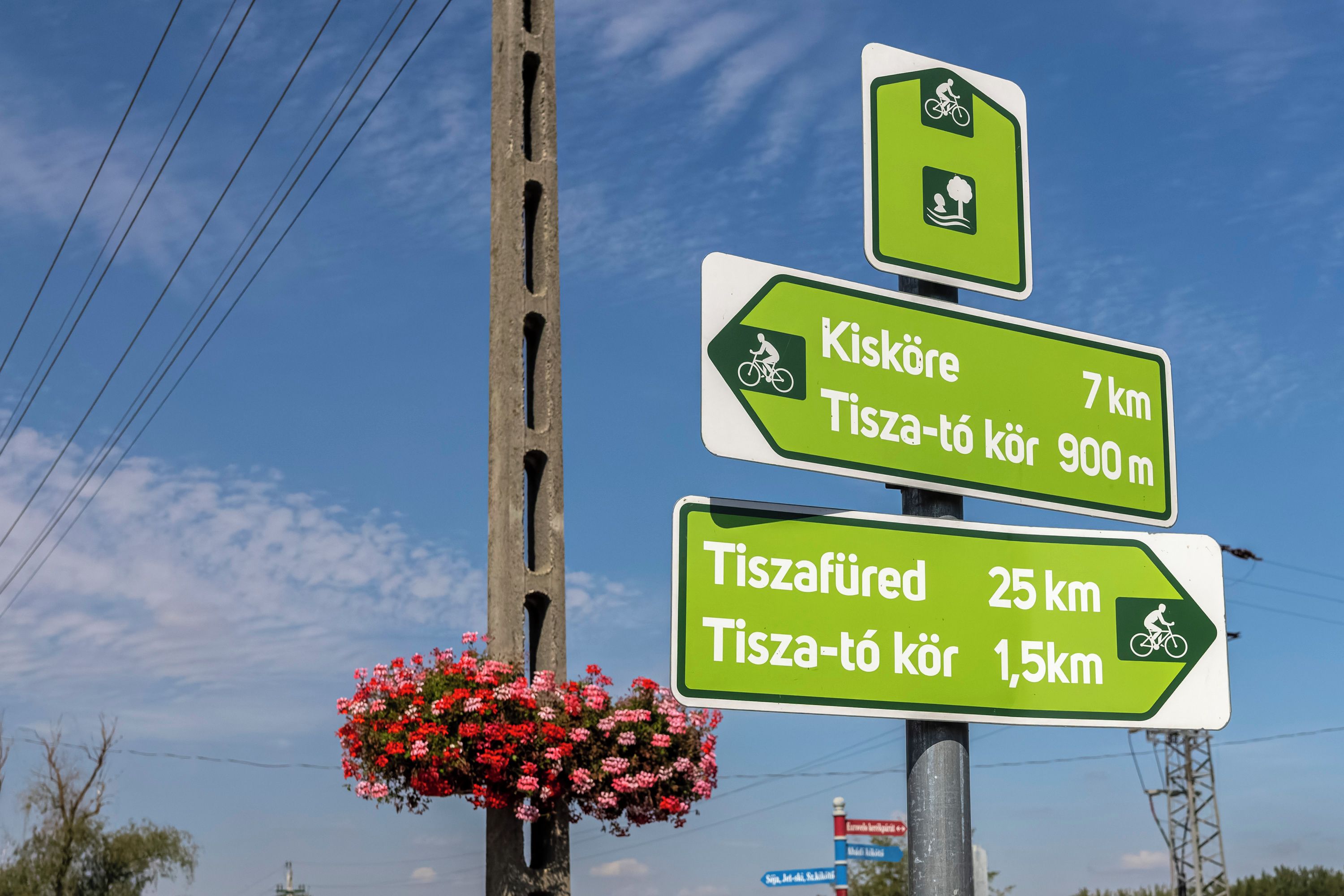 Panneaux indicateurs indiquant les distances vers Kis-Kore et Tisza-Tore, ainsi qu’un pot de fleurs fixé à un poteau.