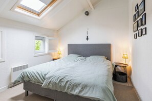 A bedroom with a skylight, a bed with a grey headboard, two bedside lamps, a window with blinds, and a wall-mounted picture frame.