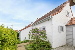 A white house with a red-tiled roof, a small window with shutters, and a garden with green foliage and purple flowers. - Sea Side Village 91* (De Haan)