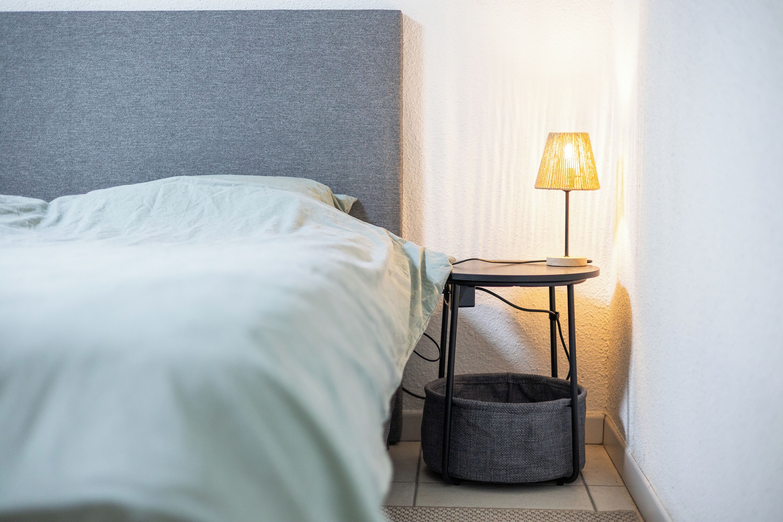 A neatly made bed with a headboard, a small table with a lamp, and a waste basket.