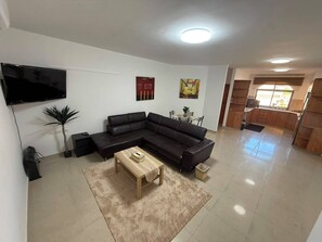 Una sala de estar con un sofá modular, un televisor de pantalla plana, una mesa de centro y un comedor con sillas.