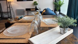 Une table à manger dressée avec de la vaisselle en verre transparent et des assiettes, une plante en pot, et une vue sur un paysage urbain par la fenêtre.