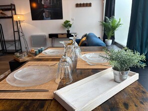 A dining table set with clear glassware and plates, a potted plant, and a view of a cityscape through a window. - Duplex lumineux centre-ville, tout équipé, parking (Le Mans)