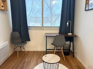 Ein Zimmer mit Schreibtisch, zwei Stühlen, einem kleinen runden Tisch, einer Heizung und einem fenster mit Vorhängen.