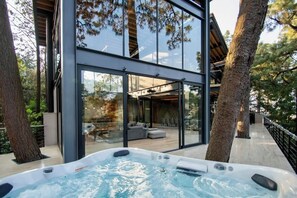 A modern outdoor hot tub area with glass walls, a wooden deck, and a view of the interior.