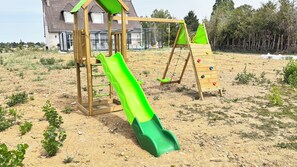 Eine hölzerne Spielkonstruktion mit Rutsche und Kletterbereich, eingebettet in eine sandige Fläche mit vereinzelter Vegetation und einem Haus im Hintergrund.