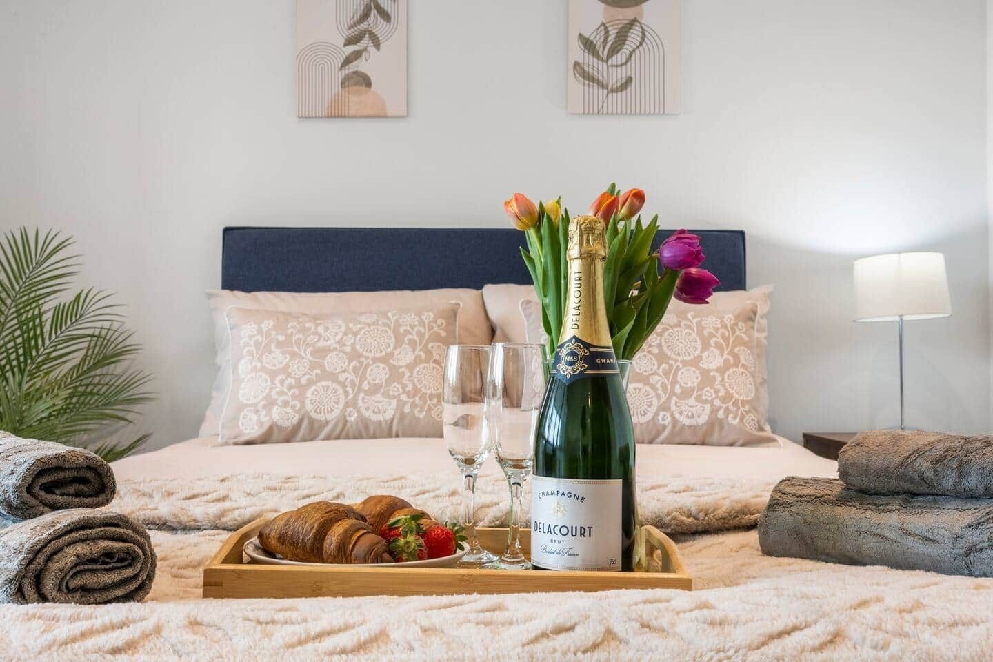 A neatly made bed with a blue headboard, a bottle of champagne, a tray with croissants and strawberries, and a vase with tulips.