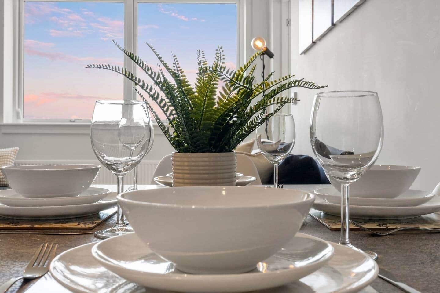 A dining table set with white plates, bowls, and wine glasses, with a fern plant centerpiece and a lit bulb on the wall.
