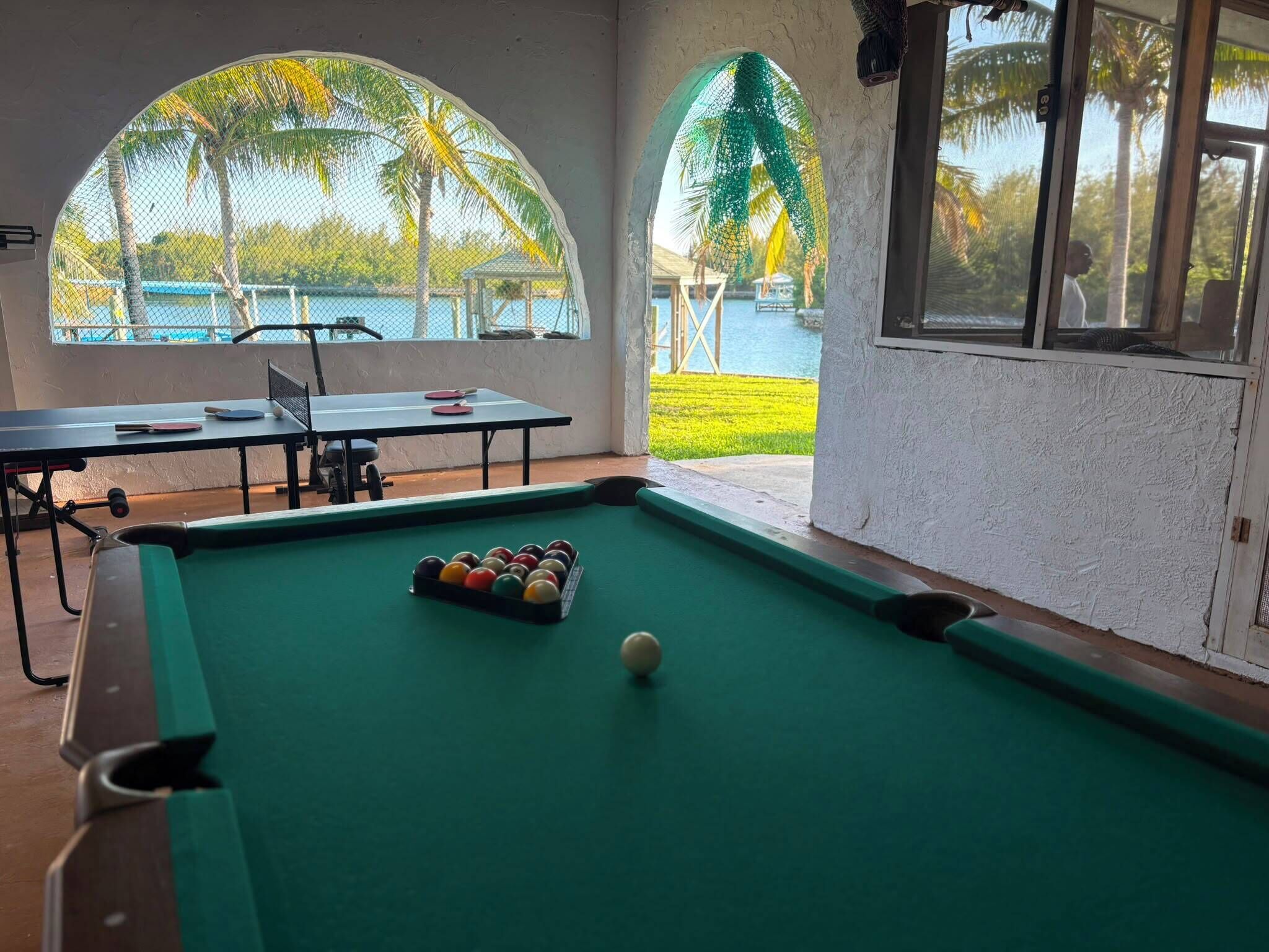 A pool table with balls, a ping pong table, and a view of a pool area with palm trees.