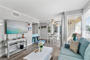 Un salon moderne doté d’un téléviseur à écran plat, d’une table basse ornée de fleurs, d’un canapé et d’un coin repas avec des chaises blanches.